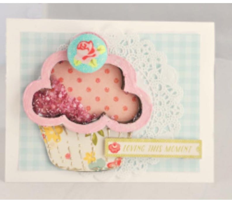 May include: A pink and white cupcake with a pink and white polka dot pattern and a blue and white flower on top. The cupcake is on a white background with a blue and white gingham pattern. The text "Loving this moment" is on a yellow banner.