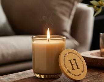 Puede incluir: Una vela perfumada encendida en un tarro de cristal transparente con una tapa de madera con la letra "H". La vela está sobre una mesa de madera, con una vela encendida en un votivo de cristal y un jarrón de flores en el fondo. La vela es beige.