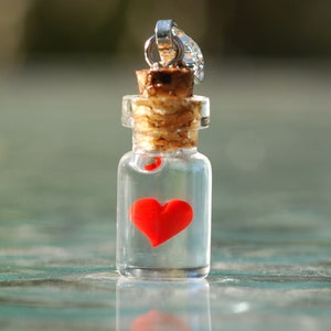 Könnte beinhalten: Ein kleiner, klarer Glasflaschenanhänger mit Korken und silberfarbener Öse. Im Inneren der Flasche befindet sich ein leuchtend rotes Herz. Der Anhänger ist wahrscheinlich ein Schmuckstück.