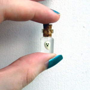 以下が含まれることがあります： コルク栓の付いた小さなガラス瓶で、小さな白い幽霊の置物が入っています。 幽霊は黒い点の目で、丸い形をしています。 瓶は2本の指で持たれています。