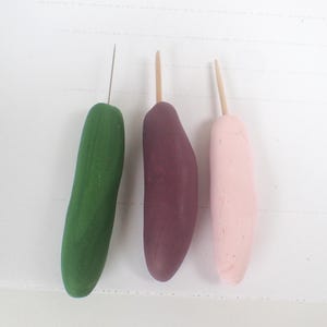 May include: Three handmade clay tools. The first is a green, cucumber-shaped tool with a metal needle. The second is a dark purple, oblong tool. The third is a light pink, oblong tool with a wooden dowel.