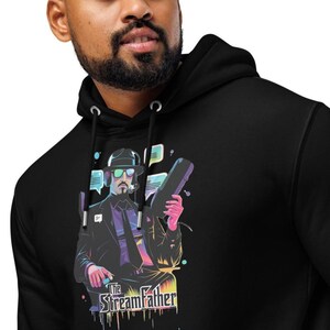 Puede incluir: Sudadera con capucha negra con un gráfico colorido de un hombre con traje, sombrero y gafas de sol, sosteniendo un dispositivo. El texto "The StreamFather" está impreso debajo. La sudadera tiene una capucha con cordón.