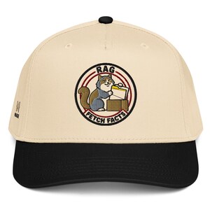 Puede incluir: Una gorra de béisbol beige con visera negra y un parche bordado circular en la parte delantera. El parche muestra un gato sosteniendo una carpeta amarilla dentro de una caja, rodeado por el texto RAG FETCH FACTS en un borde circular rojo y negro.