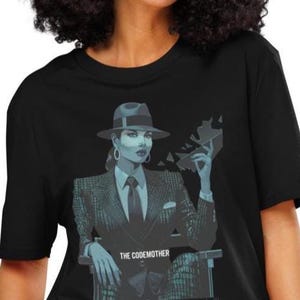 May include: Black t-shirt with a graphic of a woman in a suit and fedora holding a data chip. The text "THE CODEMOTHER" is below the image. The woman has a serious expression and is wearing a tie and earrings.