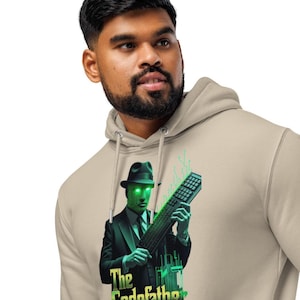 Peut inclure: Sweat à capuche beige avec un graphisme d'un homme en costume et fedora tenant un clavier, avec des lignes numériques vertes et le texte "The Codefather". Le design a un thème tech et mafia.