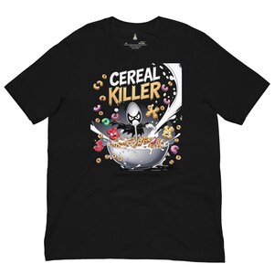 May include: A black short-sleeve t-shirt featuring a graphic print of a bowl of cereal with a spoon character wearing a black cape. The text CEREAL KILLER is printed above the bowl in yellow and white, with milk splashing and colorful cereal pieces.