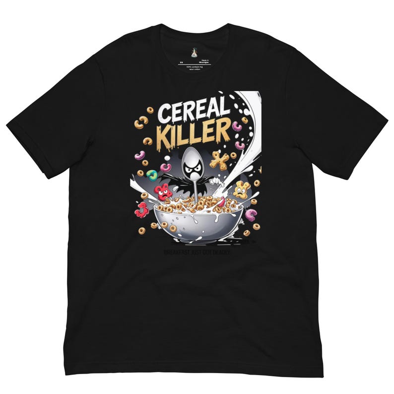 May include: A black short-sleeve t-shirt featuring a graphic print of a bowl of cereal with a spoon character wearing a black cape. The text CEREAL KILLER is printed above the bowl in yellow and white, with milk splashing and colorful cereal pieces.
