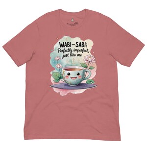 May include: A dusty rose short-sleeve t-shirt featuring a graphic of a smiling teacup with gold cracks, surrounded by lotus flowers and watercolor splashes. The text reads: WABI-SABI: Perfectly imperfect, just like me. The shirt has a crew neckline.