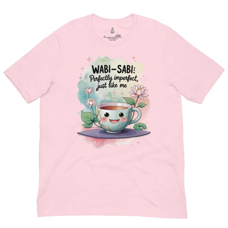 May include: A light pink t-shirt featuring a graphic of a smiling teacup with gold cracks, surrounded by lotus flowers and watercolor splashes. The text reads: WABI-SABI: Perfectly imperfect, just like me. The design includes small star accents.