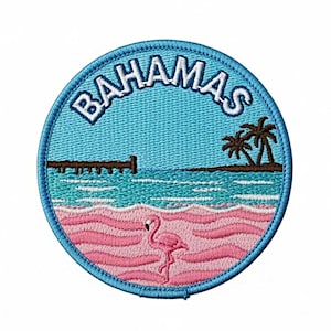 May include: A circular embroidered patch featuring a tropical scene with a pink flamingo standing on a pink beach, turquoise ocean water, a brown wooden pier, and palm trees under a light blue sky with the word BAHAMAS stitched in white letters.