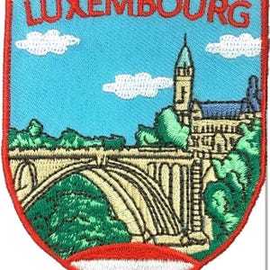 Puede incluir: Un parche bordado en forma de escudo con la palabra LUXEMBOURG en letras rojas en la parte superior. El diseño muestra un puente de piedra y un edificio contra un cielo azul claro con nubes blancas, rodeado de árboles verdes, con un motivo de bandera roja, blanca y azul en la base.
