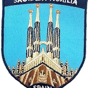 May include: A shield-shaped embroidered patch featuring a light blue background with a depiction of the Sagrada Familia in tan and brown thread. The text SAGRADA FAMILIA is stitched in white at the top, and the word SPAIN is stitched in white at the bottom.