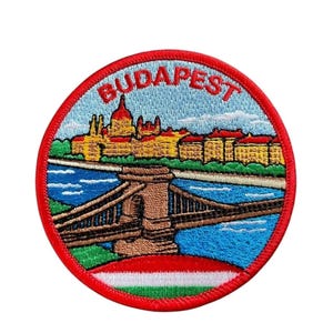 May include: A circular embroidered patch featuring a colorful scene of Budapest. The design includes the city skyline, the Chain Bridge over a blue river, and the word BUDAPEST in red letters at the top, all bordered by a thick red stitched edge.
