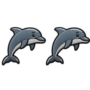 May include: Two identical embroidered patches featuring a cartoon dolphin in shades of grey and white. Each dolphin is depicted in a leaping pose with a black stitched border, a smooth grey upper body, and a white underbelly with a friendly expression.
