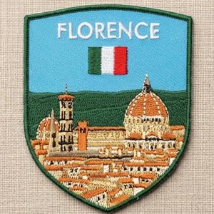 May include: A shield-shaped embroidered patch featuring a light blue sky with the text FLORENCE in white letters above a small Italian flag. The lower portion depicts a detailed cityscape with terracotta rooftops and a large domed cathedral.