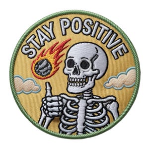 Op de afbeelding: Ronde geborduurde patch met de tekst "STAY POSITIVE". Het ontwerp toont een skelet dat een duim omhoog steekt, een vurige meteoor en wolken, alles op een gele achtergrond met een groene rand.