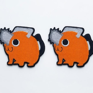 May include: Two embroidered patches featuring a stylized orange creature with a gray chainsaw blade protruding from its head. The character has a large black eye, a small mouth, and a black outline, set against a plain white background.