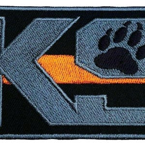 Può includere: Toppa ricamata con le lettere "K9" in grigio, una striscia arancione e un'impronta di zampa nera. La toppa ha un bordo nero e una forma rettangolare. Il disegno è cucito su uno sfondo nero.