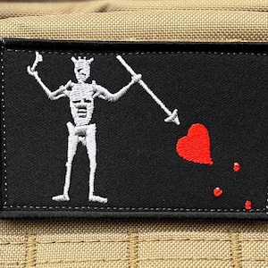 Può includere: Toppa rettangolare nera con una figura di scheletro bianco che tiene una lancia, un cuore rosso e goccioline. La toppa ha un bordo ricamato nero ed è posta su uno sfondo beige. Il design ricorda una bandiera pirata.
