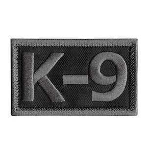 Op de afbeelding: Een rechthoekige patch met de geborduurde tekst "K-9" in grijs op een zwarte achtergrond. De patch heeft een grijze rand en is waarschijnlijk bedoeld om op kleding of uitrusting te worden genaaid of bevestigd.