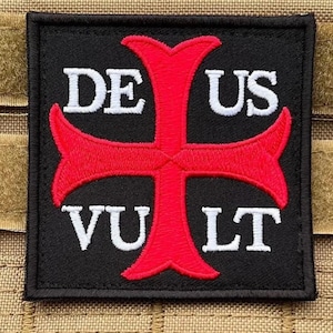 Peut inclure: Un écusson carré noir avec une croix rouge et des lettres blanches. Les mots "DE US VULT" sont affichés sur l'écusson. L'écusson est probablement un emblème ou une insigne.