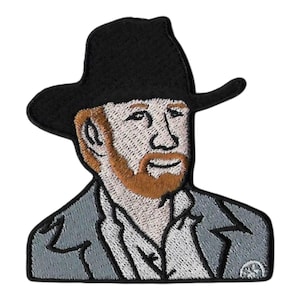 May include: An embroidered patch featuring a person wearing a black cowboy hat, a light gray collared jacket, and a white shirt. The figure has light skin, reddish-brown hair, and a matching beard, depicted in a stylized, stitched portrait format.