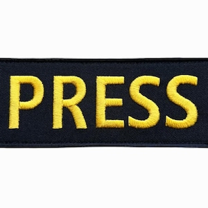 Puede incluir: Un parche rectangular azul marino con la palabra "PRESS" bordada en hilo amarillo. Las letras amarillas destacan sobre el fondo oscuro. El parche está diseñado para ser aplicado en ropa o equipo.