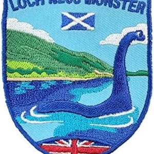 May include: A shield-shaped embroidered patch featuring a light blue background with the text LOCH NESS MONSTER at the top. It depicts a dark blue creature in water, a green hillside, a white and blue Scottish flag, and a red, white, and blue Union Jack.