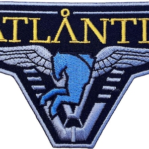 Op de afbeelding: Een donkerblauwe geborduurde patch met het woord ATLANTIS in gele hoofdletters. Onder de tekst staat een lichtblauwe afbeelding van een gevleugeld paard met gestileerde vleugels die naar buiten uitsteken, omrand door een lichtblauwe stikselrand.