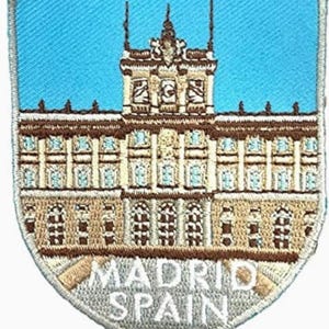 May include: A shield-shaped embroidered patch featuring a detailed depiction of a large building with brown and cream architectural elements against a bright blue sky. The text MADRID SPAIN is stitched in white capital letters at the bottom.