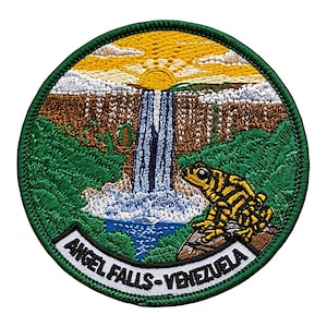 Könnte beinhalten: Ein runder bestickter Aufnäher mit einer Landschaft der Angel Falls in Venezuela, einem gelben Sonnenaufgang, weißen Wolken, braunen Klippen, grünem Laub und einem blauen Wasserfall. Ein gelb-schwarzer Frosch sitzt auf einem Felsen mit dem Text ANGEL FALLS-VENEZUELA.