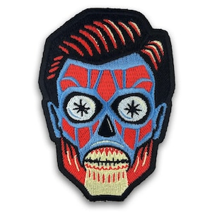 Op de afbeelding: Geborduurde patch met een gestileerd zombiegelaat. Het ontwerp omvat een zwarte omtrek, rood haar, blauwe huid en een rood en blauw gezicht met witte ogen en een grijnzende mond. Voor kleding of accessoires.