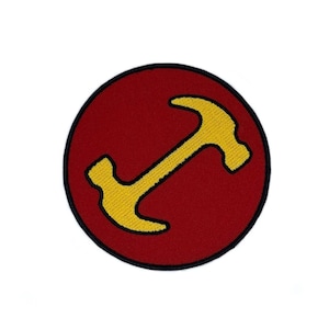 May include: A circular embroidered patch featuring a deep red background with a thick black border. Centered on the patch is a stylized yellow hammer icon positioned diagonally, with the head of the hammer pointing toward the upper right corner.