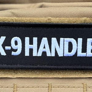 May include: A black rectangular patch with the embroidered text "K-9 HANDLER" in white. The patch is affixed to a tan fabric background, likely a tactical vest or bag. The patch has a velcro backing.