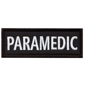 May include: A rectangular black patch with the word "PARAMEDIC" embroidered in white capital letters. The patch has a dark brown border and is likely designed to be sewn onto clothing or gear.