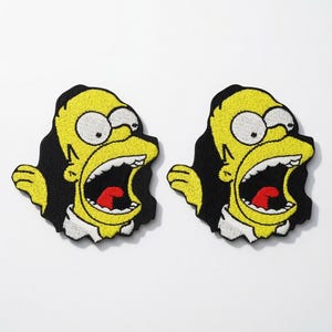 Op de afbeelding: Twee identieke geborduurde patches met een gele stripfiguur met een wijd open mond, een rode tong en witte ogen. Het personage is zwart omlijnd, geplaatst tegen een zwarte achtergrond en draagt een witte kraag aan de onderkant.