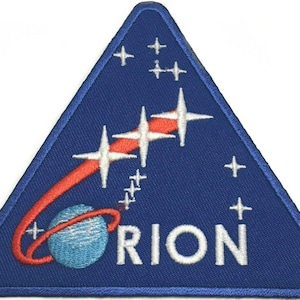 Puede incluir: Un parche bordado triangular con fondo azul oscuro. Presenta un planeta azul claro con un anillo rojo, una estela roja curva, tres grandes estrellas blancas de cuatro puntas, varias estrellas blancas pequeñas y el texto blanco ORION.