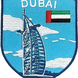 Op de afbeelding: Een schildvormige geborduurde patch met een lichtblauwe achtergrond met het Burj Al Arab-gebouw in wit en blauw, de tekst DUBAI in het wit en de vlag van de Verenigde Arabische Emiraten in de kleuren rood, groen, wit en zwart.