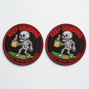 May include: Two circular embroidered patches featuring a white skeleton walking on a green patch of grass while holding a tan coffee mug. The black background is bordered in red with the text KEEP GRINDING EVEN IF YOU'RE DEAD INSIDE.