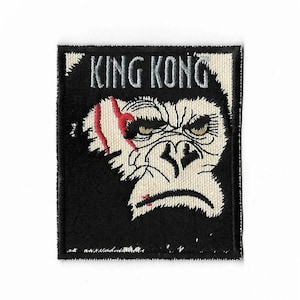 Peut inclure: Un écusson brodé rectangulaire représentant une illustration en noir et blanc d'un visage de gorille avec une cicatrice rouge sur l'œil droit. Le texte KING KONG est brodé en gris en haut. Le motif est sur un fond noir.