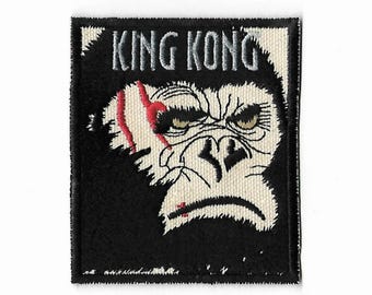 Può includere: Una toppa ricamata rettangolare con un'illustrazione in bianco e nero di un volto di gorilla con una cicatrice rossa sopra l'occhio destro. Il testo KING KONG è ricamato in grigio nella parte superiore. Il design è su sfondo nero.