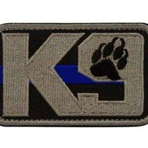Puede incluir: Parche rectangular con borde negro, con las letras "K9" en gris, una franja azul y una huella de pata negra. El parche está diseñado con detalles bordados y un aspecto texturizado.