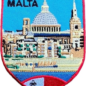 Może przedstawiać: Naszywka w kształcie tarczy z jasnoniebieskim tłem i czarnym napisem MALTA. Wzór przedstawia szczegółową scenę architektoniczną z centralną kopułą, wysoką wieżą oraz małą flagą Malty umieszczoną w dolnej części naszywki.