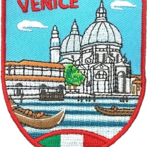 Puede incluir: Un parche bordado en forma de escudo con un cielo azul claro, nubes blancas y el texto rojo VENICE. Representa una catedral blanca con cúpula, edificios marrones y dos góndolas marrones sobre agua azul, con un borde rojo y la bandera de Italia.