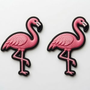 Könnte beinhalten: Zwei gestickte Aufnäher, die jeweils einen auf einem Bein stehenden rosa Flamingo zeigen. Die Flamingos haben eine texturierte rosa Fadenstickerei, schwarze Umrandungen sowie schwarz-weiße Schnäbel. Die Motive sind identisch im Profil.