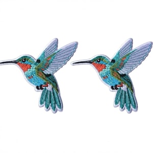 Peut inclure: Deux écussons brodés identiques représentant un colibri en vol. L'oiseau possède une gorge orange vif, une poitrine bleu clair et des ailes vertes avec des accents blancs. Chaque écusson présente un fin bord blanc et un long bec noir pointu.