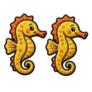 Op de afbeelding: Twee identieke geborduurde patches in de vorm van zeepaardjes. Elk zeepaardje is felgeel met oranje stippen, een oranje rugvin en een gekrulde staart. De patches zijn voorzien van een dikke zwarte rand die de vorm omlijnt.