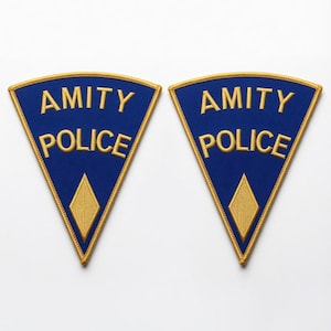 May include: Two triangular embroidered patches with a blue background and gold border. The text AMITY POLICE is stitched in gold thread across the center of each patch, with a gold diamond shape positioned below the text near the bottom point.