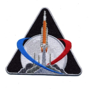 Op de afbeelding: Een driehoekige geborduurde patch met een raket die opstijgt vanaf een grijs maanoppervlak tegen een zwarte achtergrond. De raket is oranje en wit, geflankeerd door een blauwe boog links en een rode boog rechts, met grijze rook aan de basis.