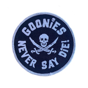Può includere: Toppa rotonda blu navy con testo ricamato bianco e un disegno di teschio e ossa incrociate. Il testo recita "GOONIES NEVER SAY DIE!" in una disposizione circolare. La toppa ha un bordo bianco cucito.